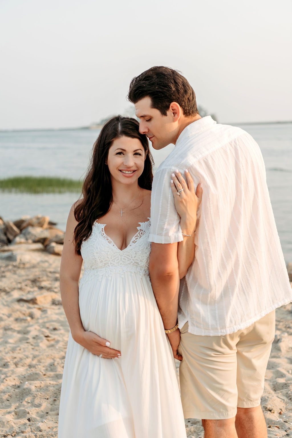 A mom to be in a white maternity gown stands on a beach with her husband nuzzling her head