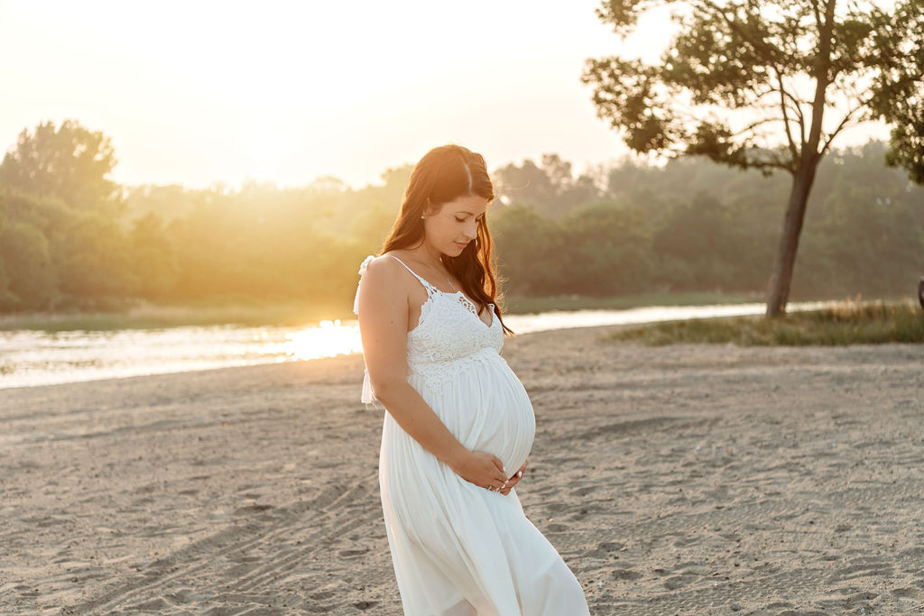 A mother to be smiles down to her bump while standing on a beach at sunset in a white maternity gown after visiting a birthing center stamford ct