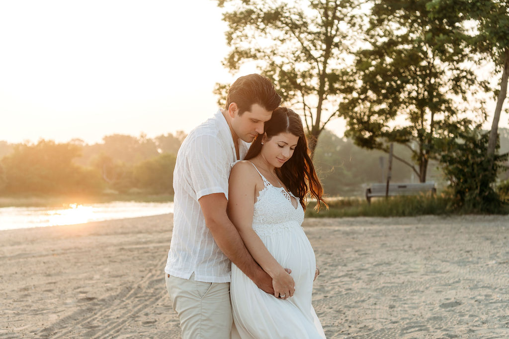 A mother to be in a white maternity gown stands on a beach at sunset while being hugged from behind by her husband after visiting a birthing center stamford ct