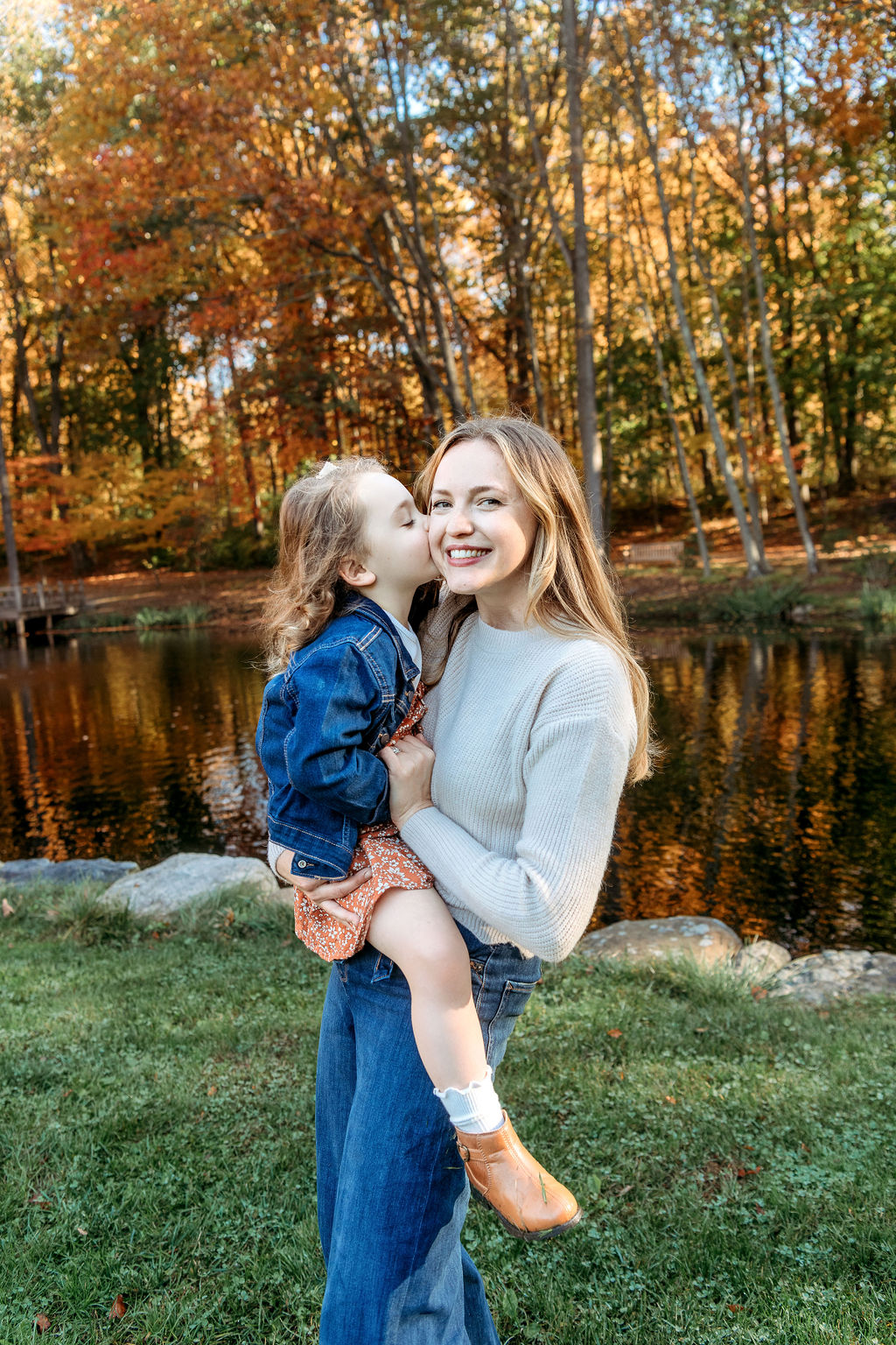 A mother in a sweater and jeans gets kissed by her toddler daughter on the cheek while standing in a riverside lawn after visiting a stamford pediatric dentist