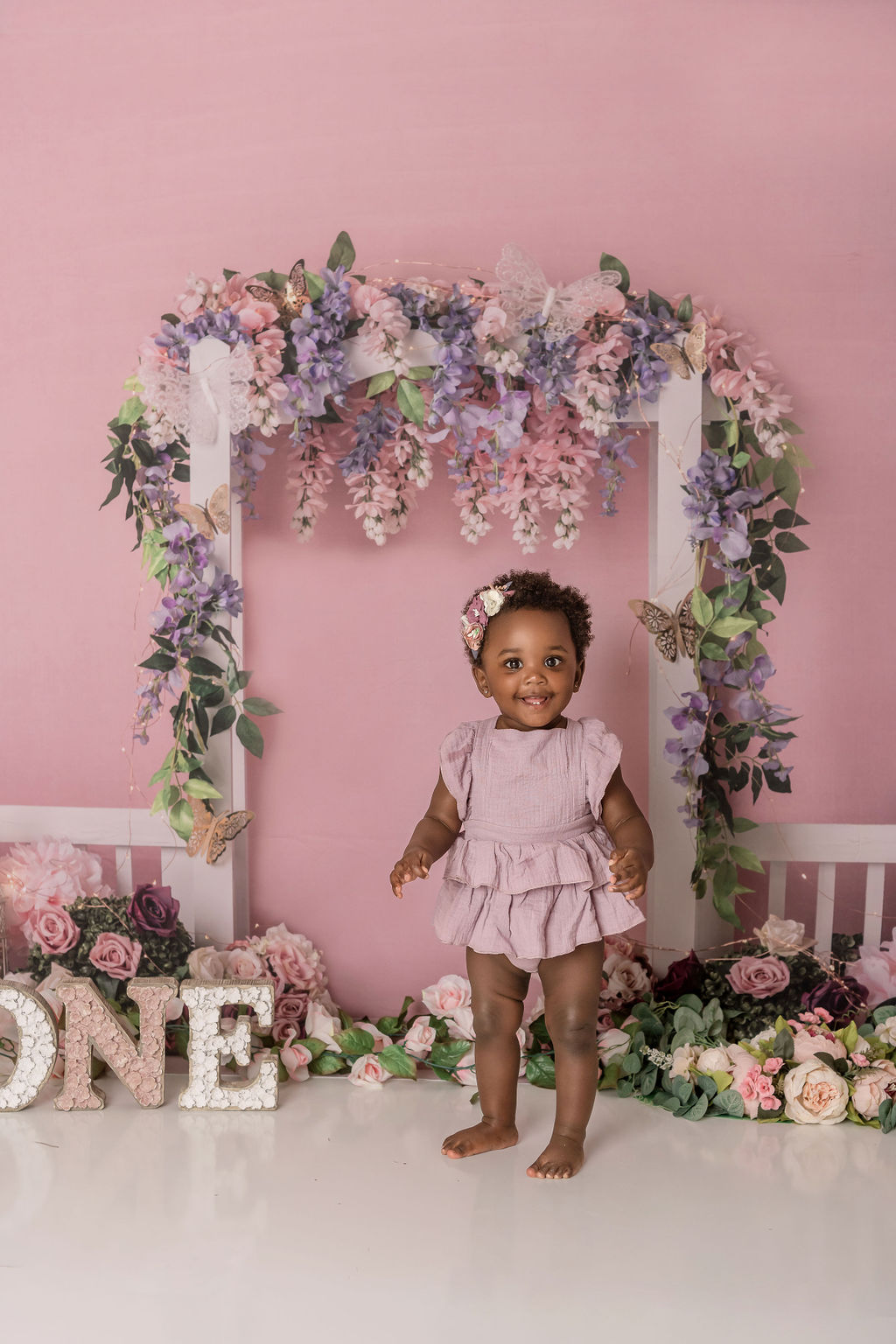 A toddler girl stands in a studio under an arbor of purple and pink flowers in a purple dress before visiting daycare stamford ct