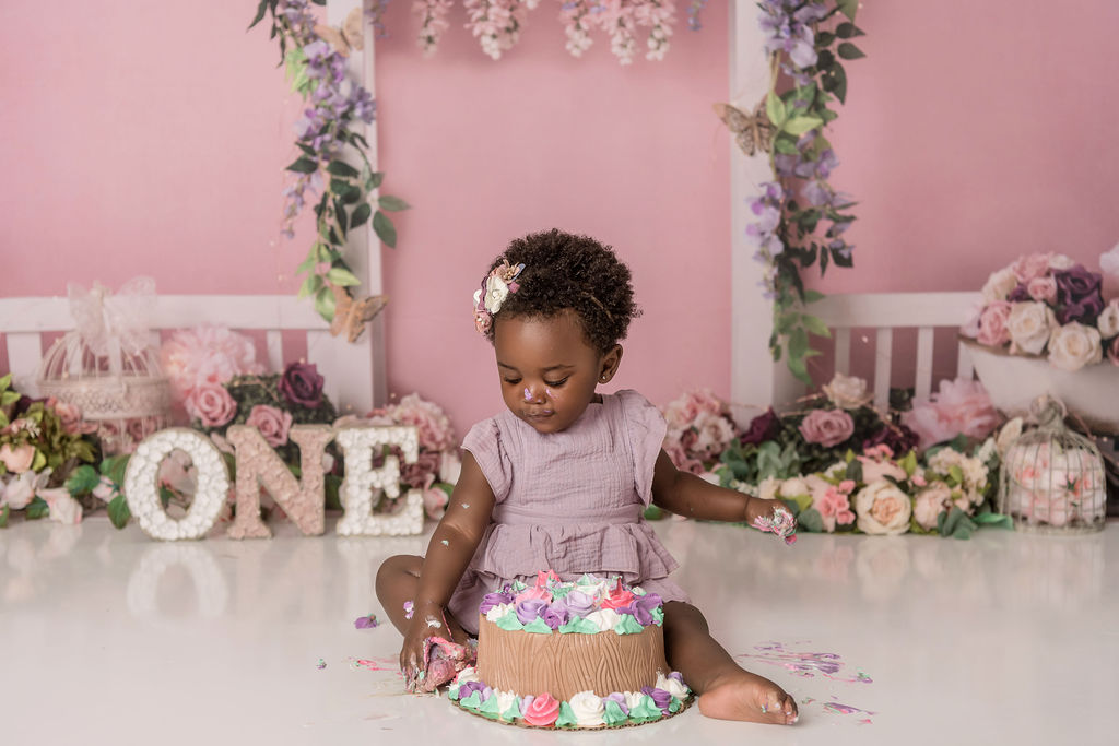 A toddler girl in a pink dress sits in a studio smashing her first birthday cake before attending daycare stamford ct