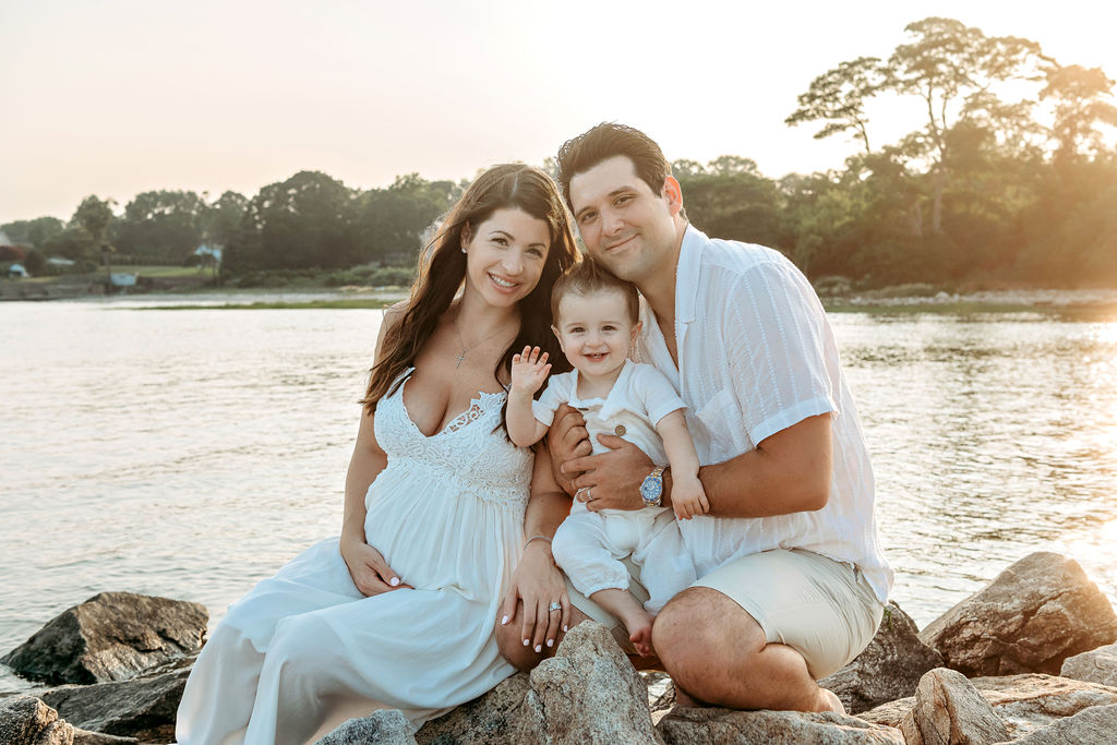 A pregnant mother in a white maternity dress sits on some rocks on the beach with her husband and toddler son at sunset after some mommy and me classes stamford ct