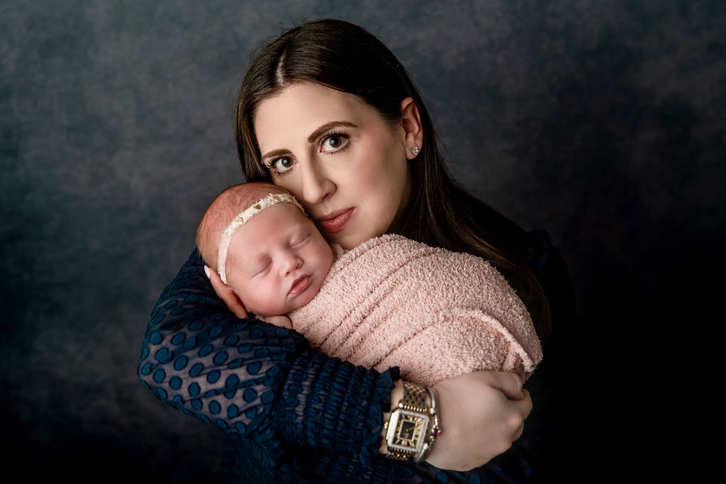 A mother in a blue dress snuggles her sleeping newborn daughter against her cheek while standing in a studio