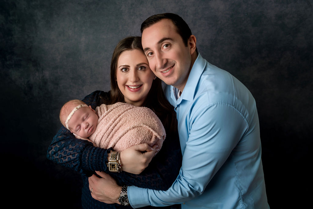 Happy new parents stand in a studio cradling their sleeping newborn baby in a pink swaddle after some parenting classes in ct