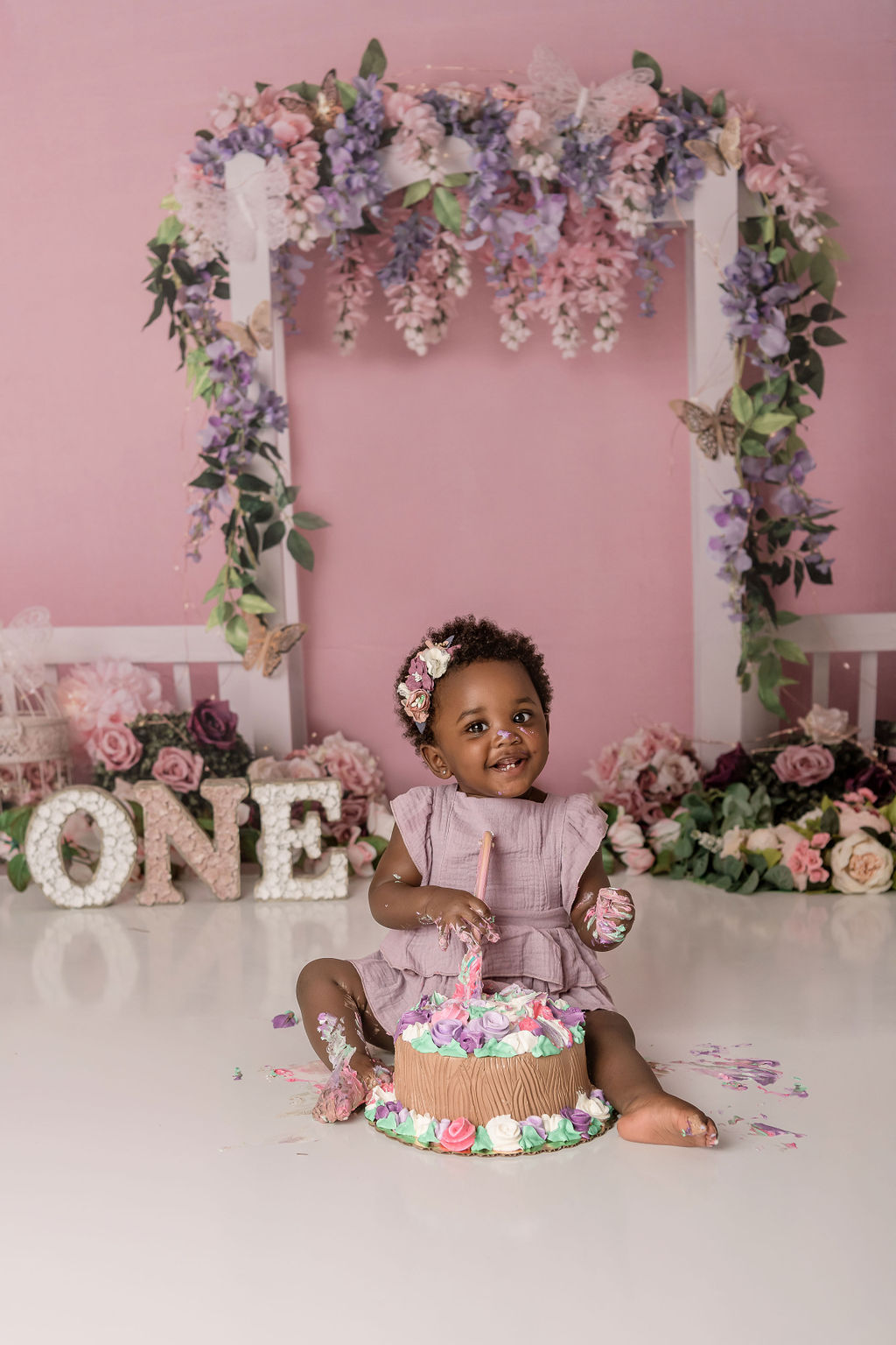 A happy toddler girl sits on a studio floor for her first birthday in a purple dress with a colorful cake
