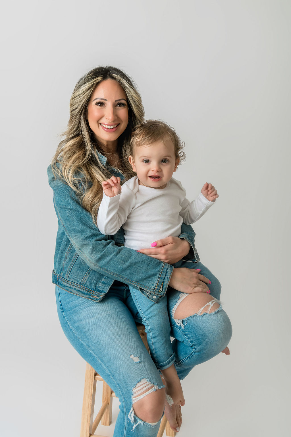 A happy mom sits on a stool in a studio wearing a denim jacket and pants with her toddler in a white shirt and jeans sitting in her lap