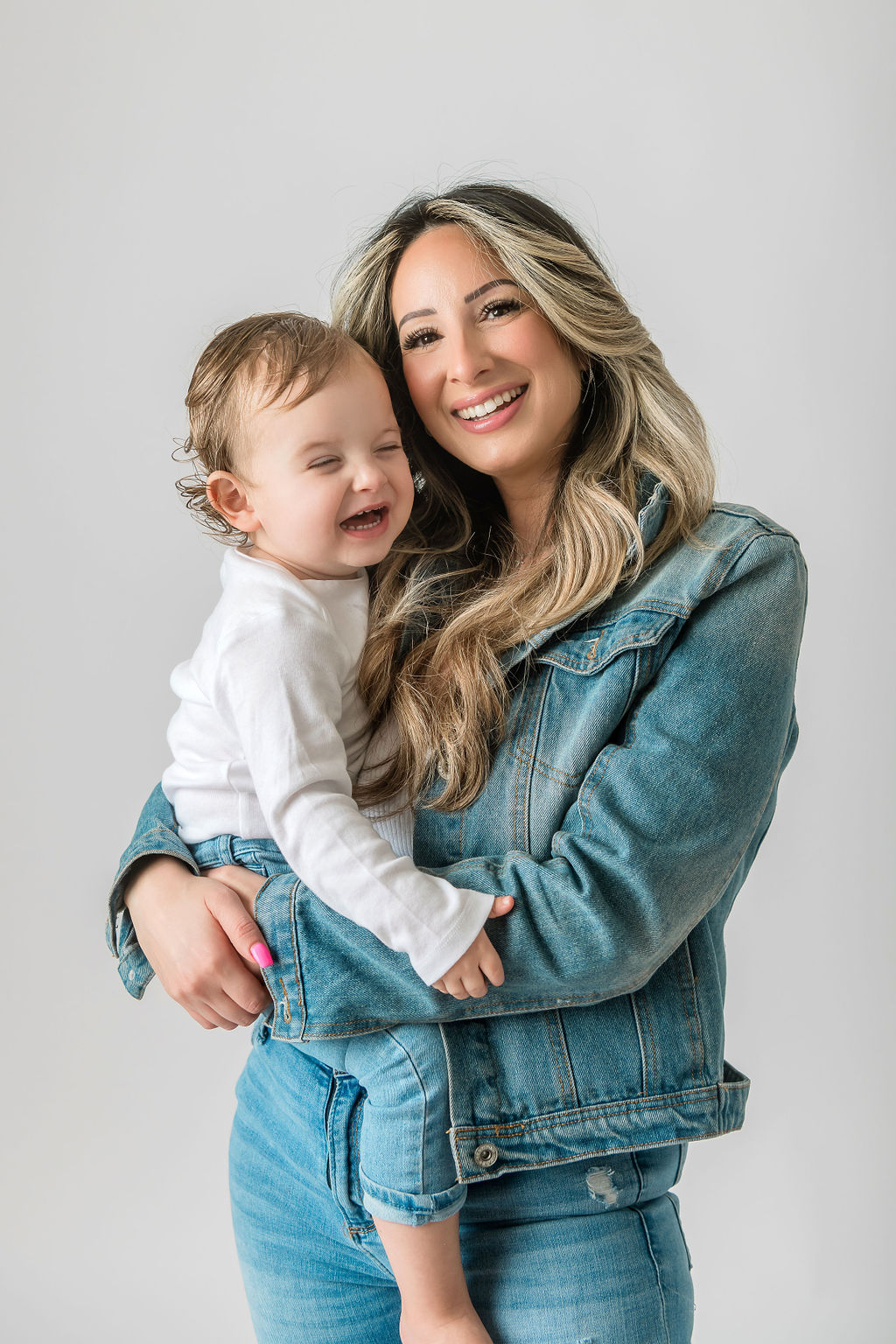 A happy mom smiles with her toddler in her arms while wearing a denim jacket and pants