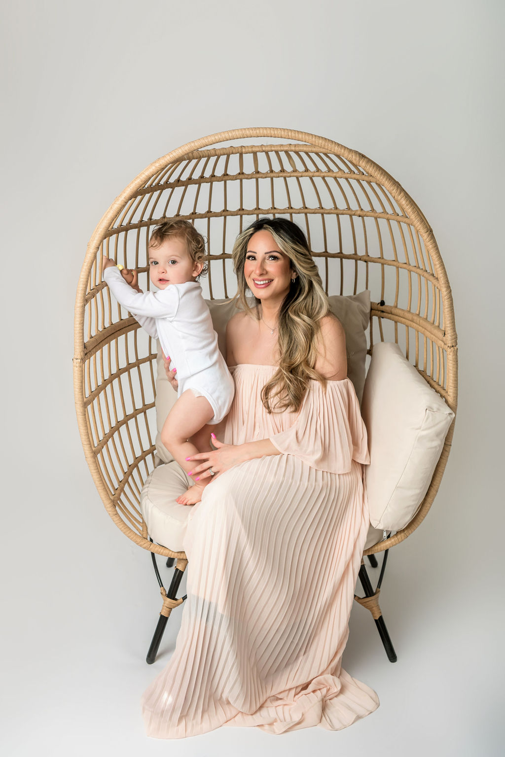 A mother in a pink dress plays in a wicker chair with her climbing toddler in a white onesie after visiting preschools stamford ct
