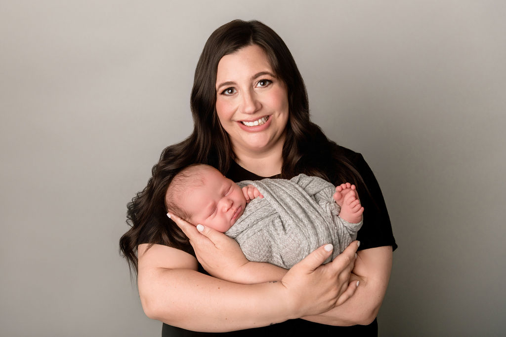 A smiling new mother in a black shirt cradles her sleeping newborn in her arms against her chest in a studio after exploring baby shower venues in Stamford, CT