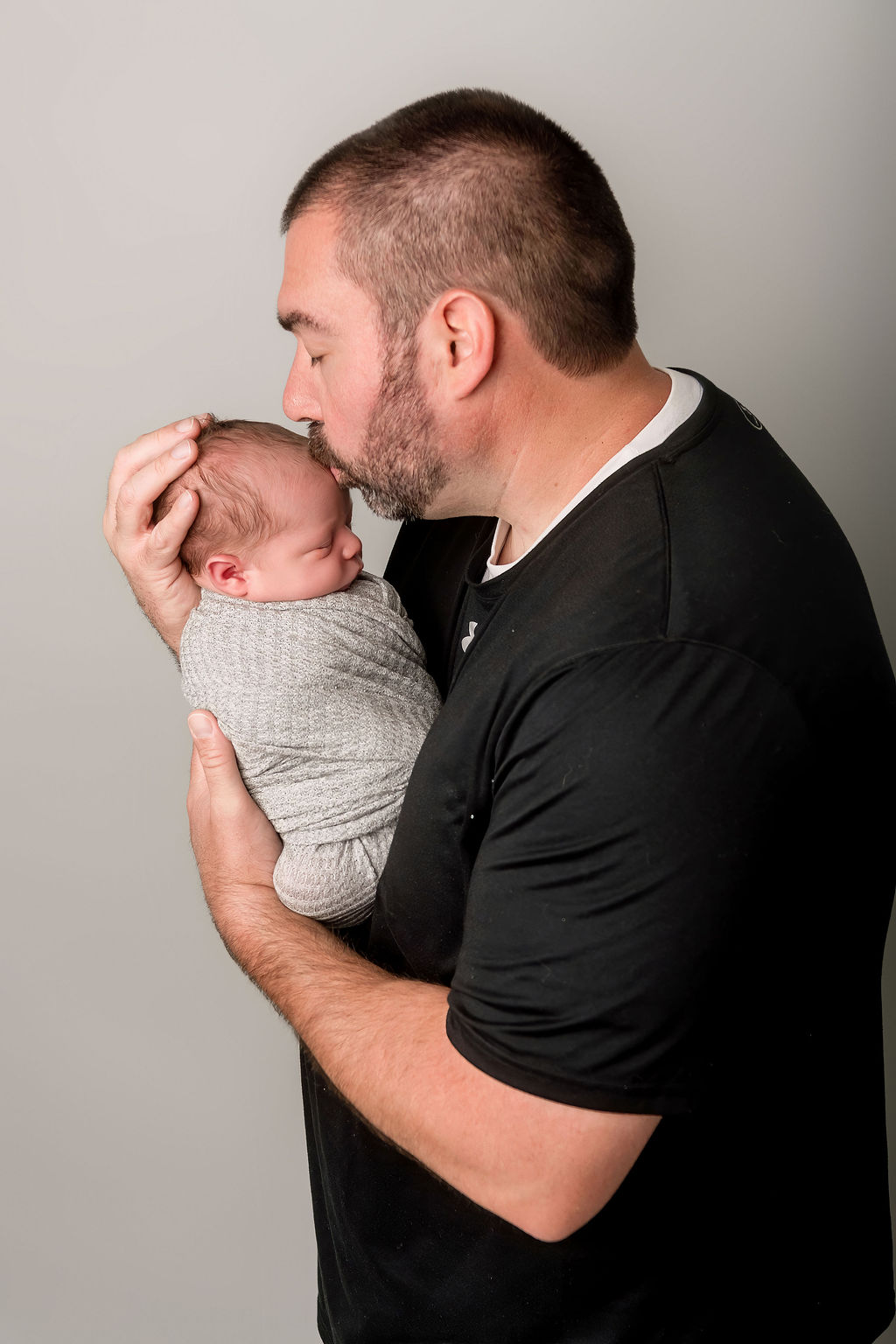 A new dad in a black shirt kisses the head of his sleeping newborn in his hands in a grey swaddle