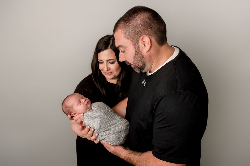 New mom and dad in black smile down at their sleeping newborn in a grey swaddle in their hands after using baby shower venues in Stamford, CT