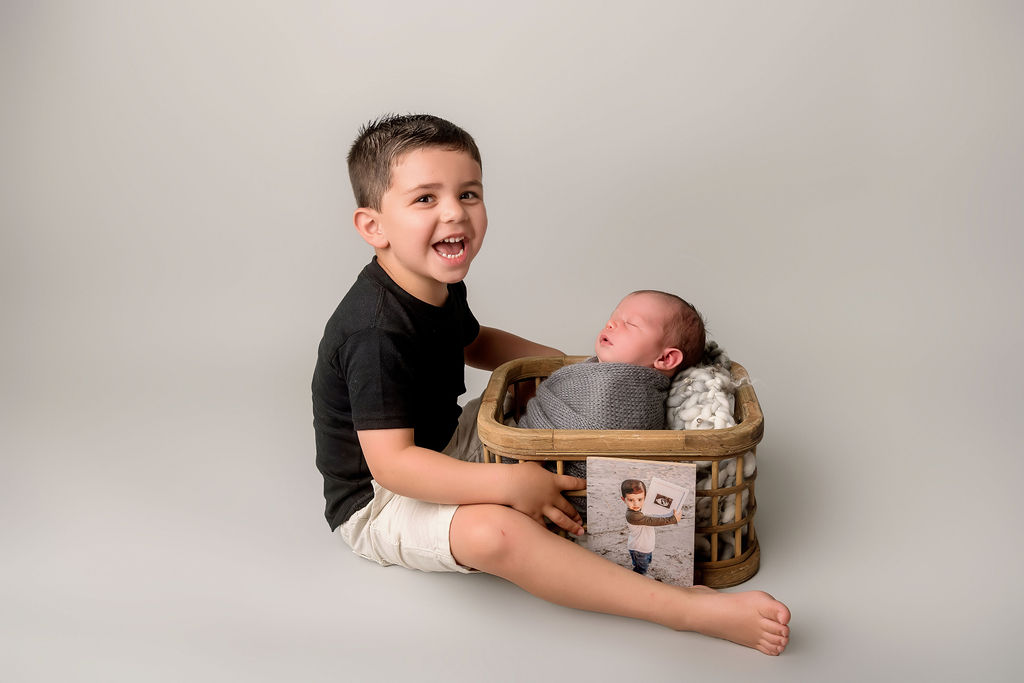 A smiling toddler sits with his newborn baby brother sleeping in a basket in his lap in a studio