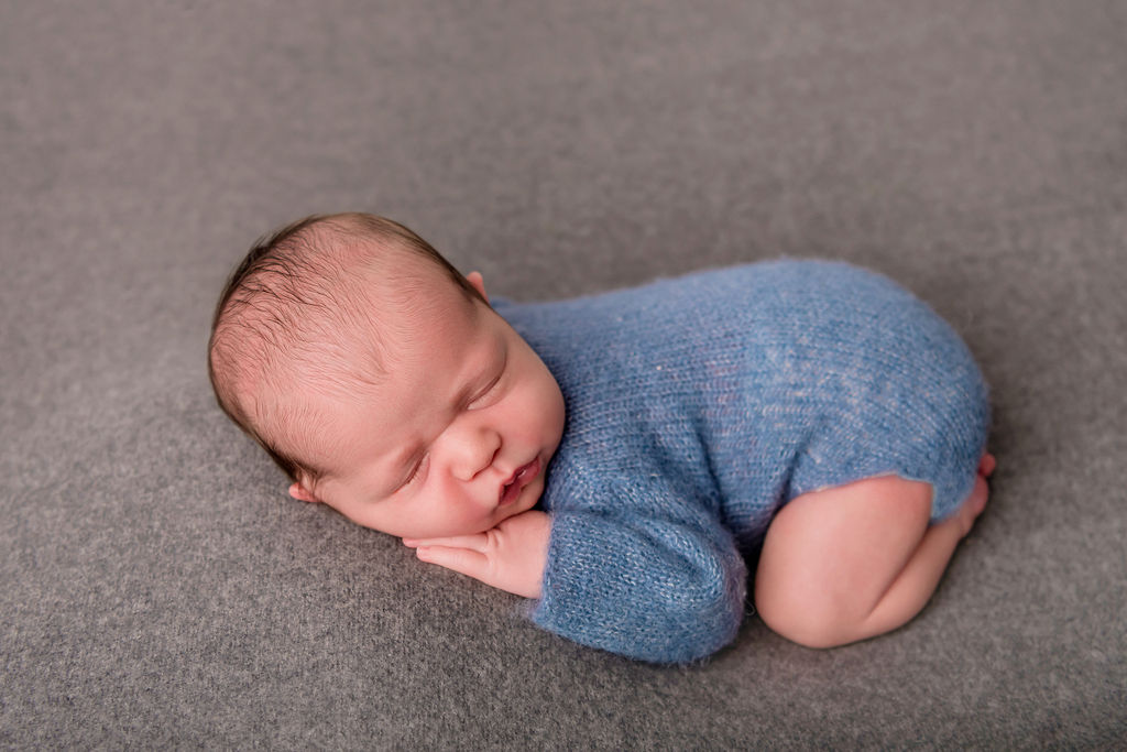A newborn baby in a blue knit onesie sleeps in froggy pose on a grey bed after visiting pediatricians in Stamford, CT