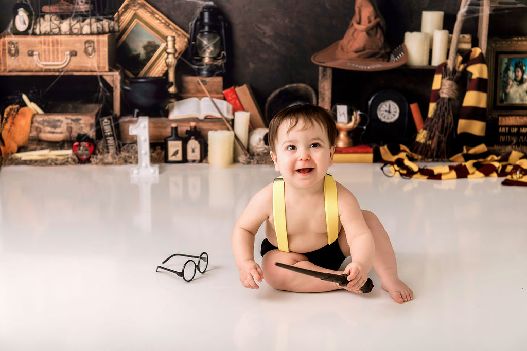 A happy baby in yellow suspenders smiles while playing with a wooden magic wand next to round glasses