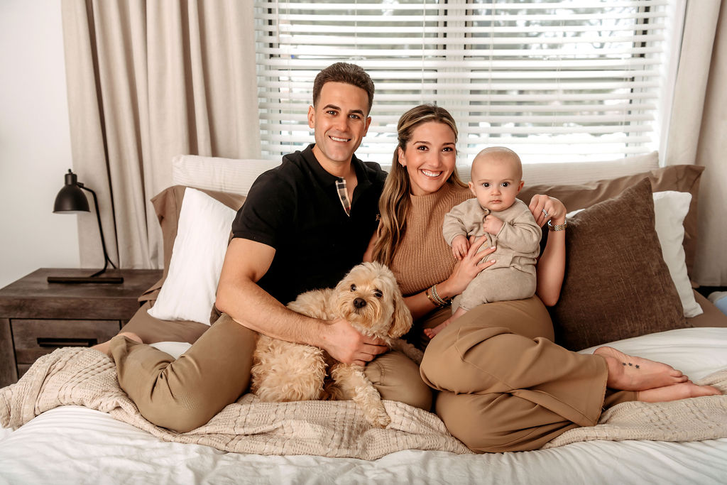 A happy new mom and dad sit on a bed with their dog and infant in their laps