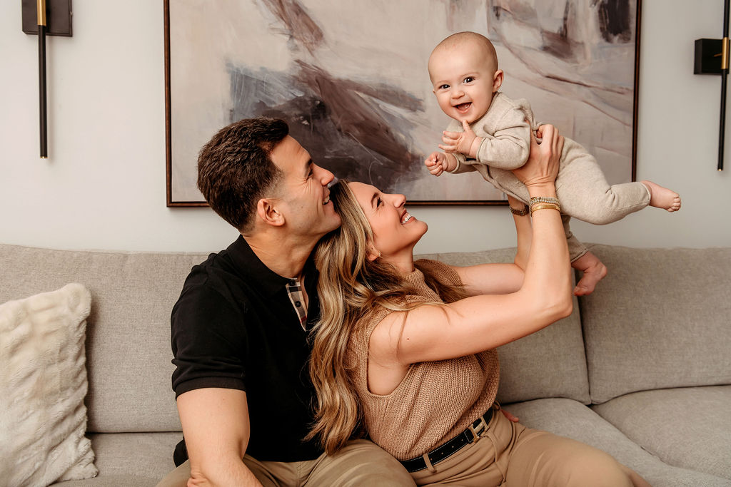 A happy baby is lifted by mom over her and dad's heads while sitting on a couch in black and tan thanks to an obgyn in Stamford, CT