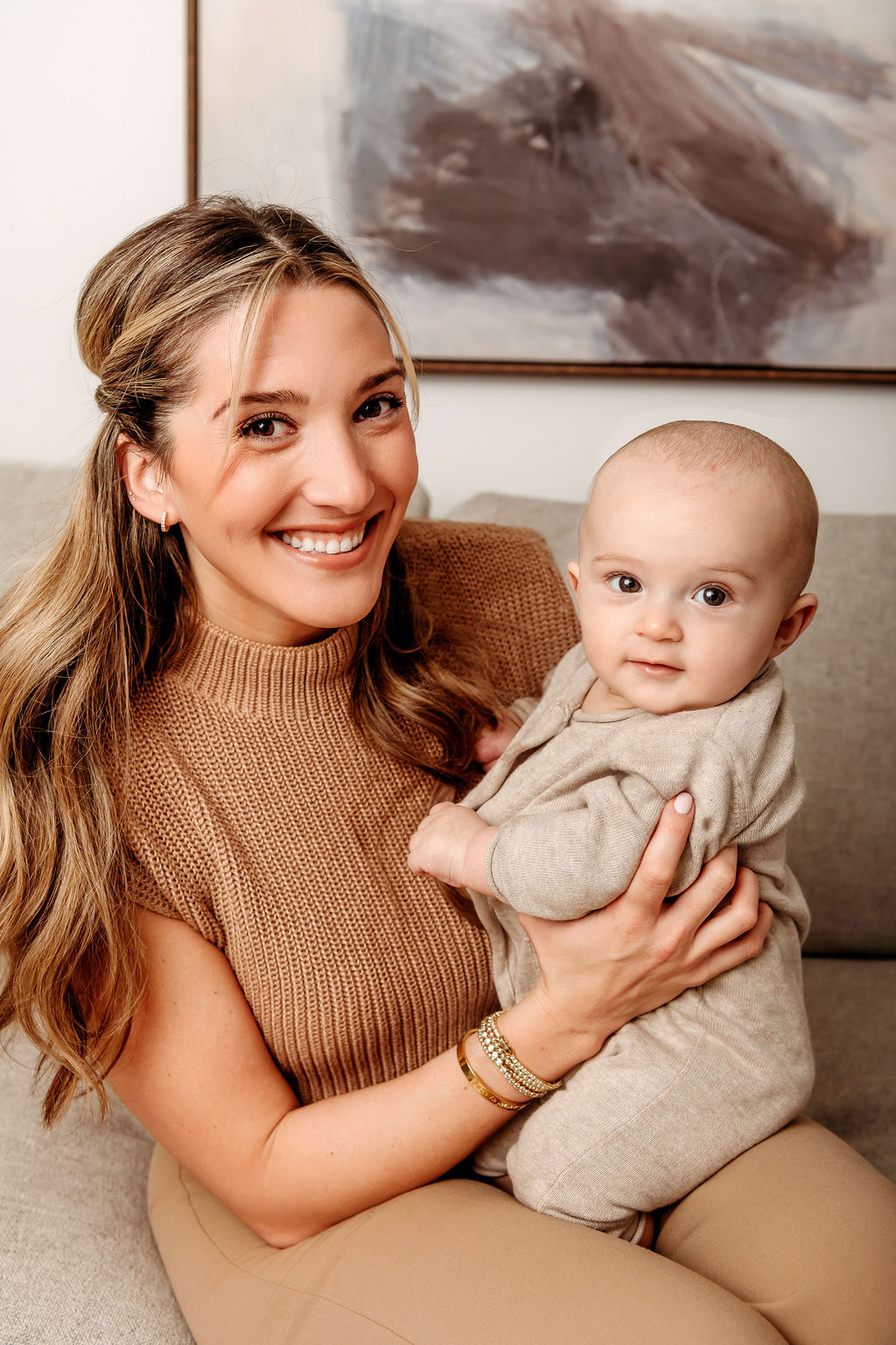A smiling new mom in a brown blouse sits on a couch with her happy baby in a tan onesie in her lap