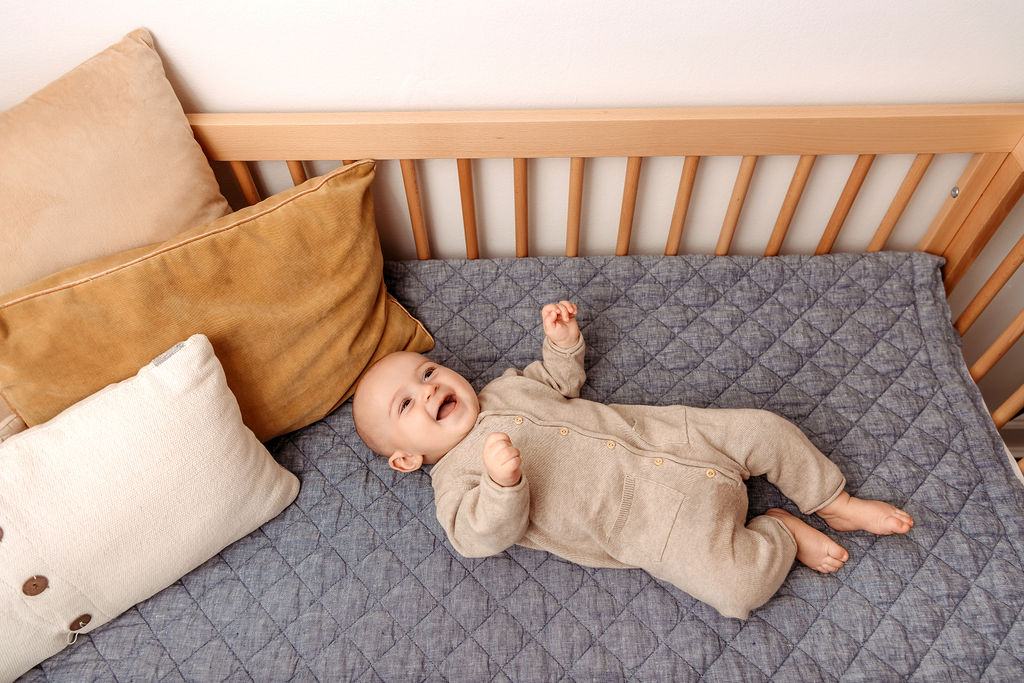 A laughing infant in a tan onesie lays in a blue crib with hands thanks to an obgyn in Stamford, CT