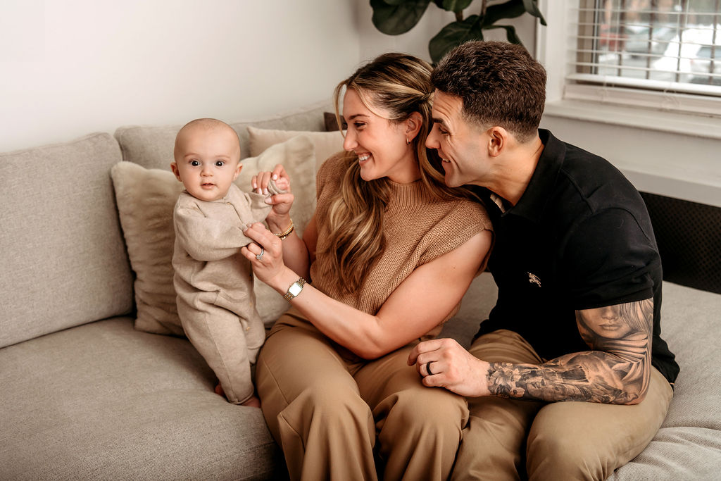 New mom and dad play with their happy baby on a couch in a tan onesie thanks to an obgyn in Stamford, CT