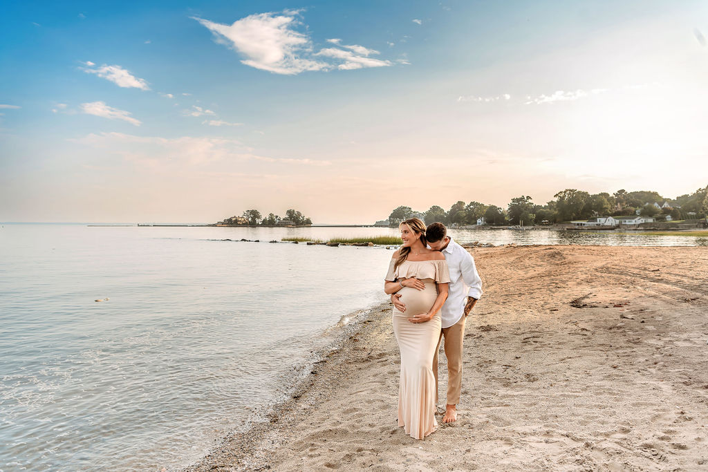 A happy expecting couple snuggle while standing on a beach at sunset holding the bump