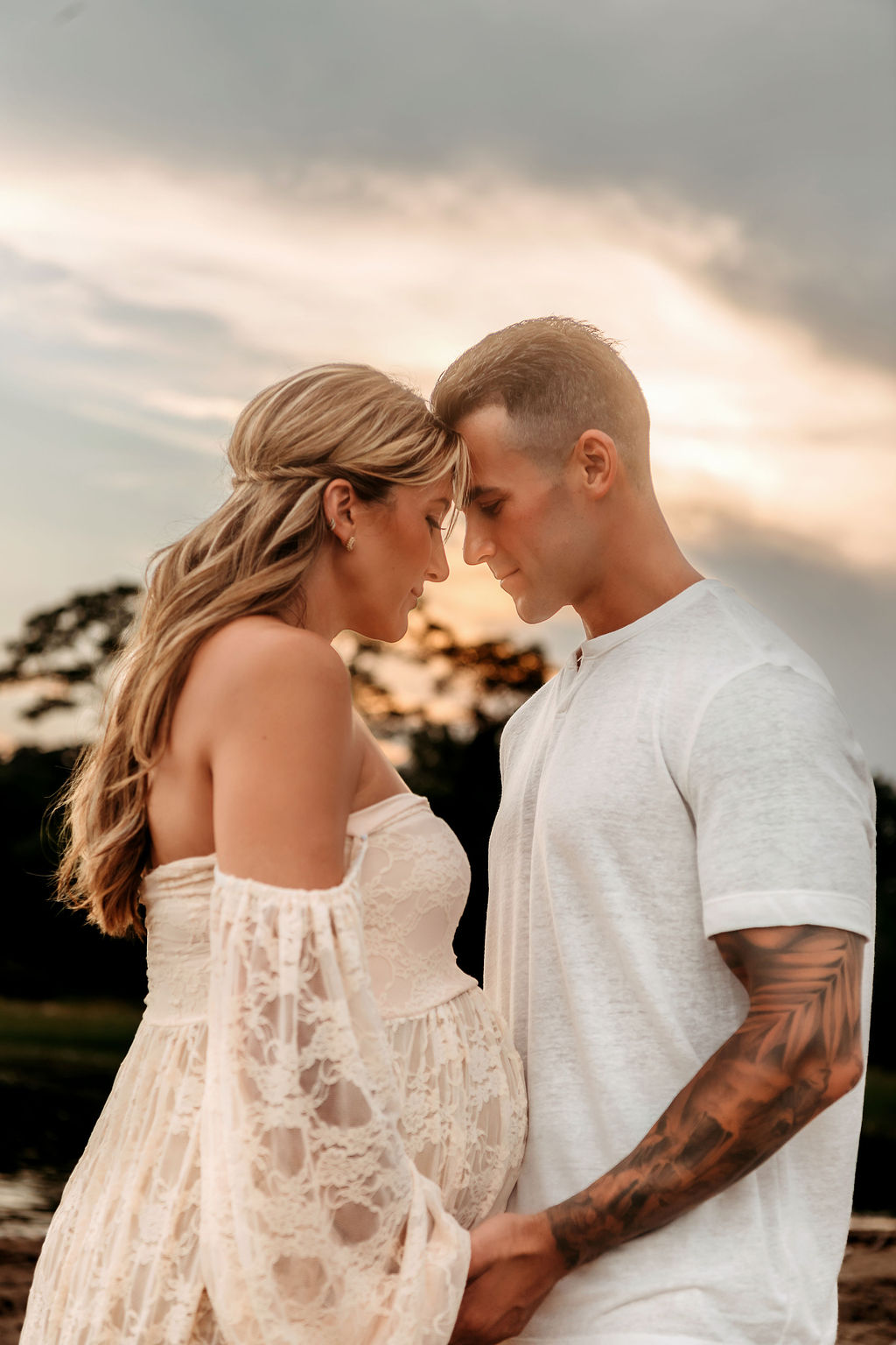 A mom to be snuggles touching foreheads with dad to be in a white lace maternity gown and white shirt at sunset