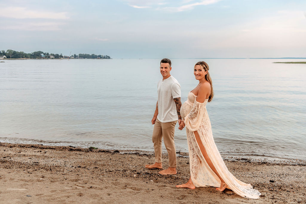 A happy pregnant couple walk on a beach in a lace maternity gown and white shirt at sunset