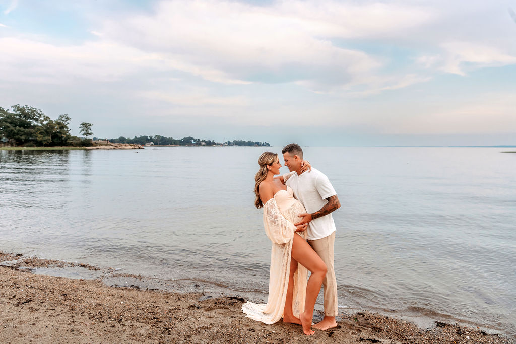 A happy pregnant couple snuggle by the water in white outfits after visiting stamford labor and delivery