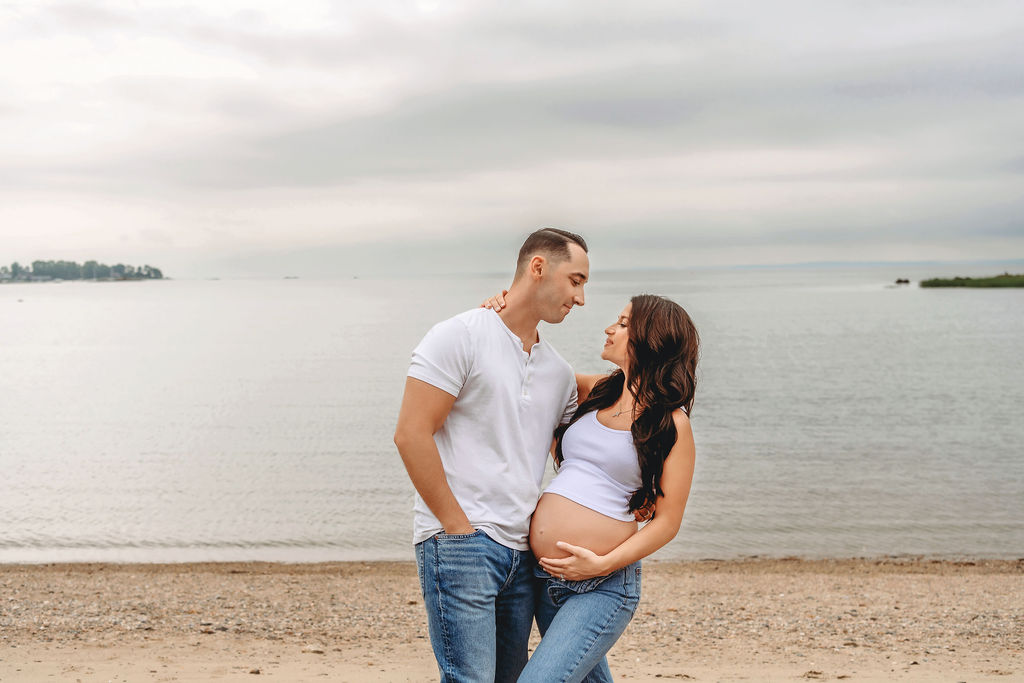 A happy pregnant couple lean in for a kiss while wearing jeans and white shirts with the bump out on a beach