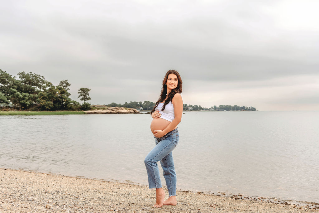A mom to be stands with hands on her bump on a beach in jeans and a white shirt at sunset