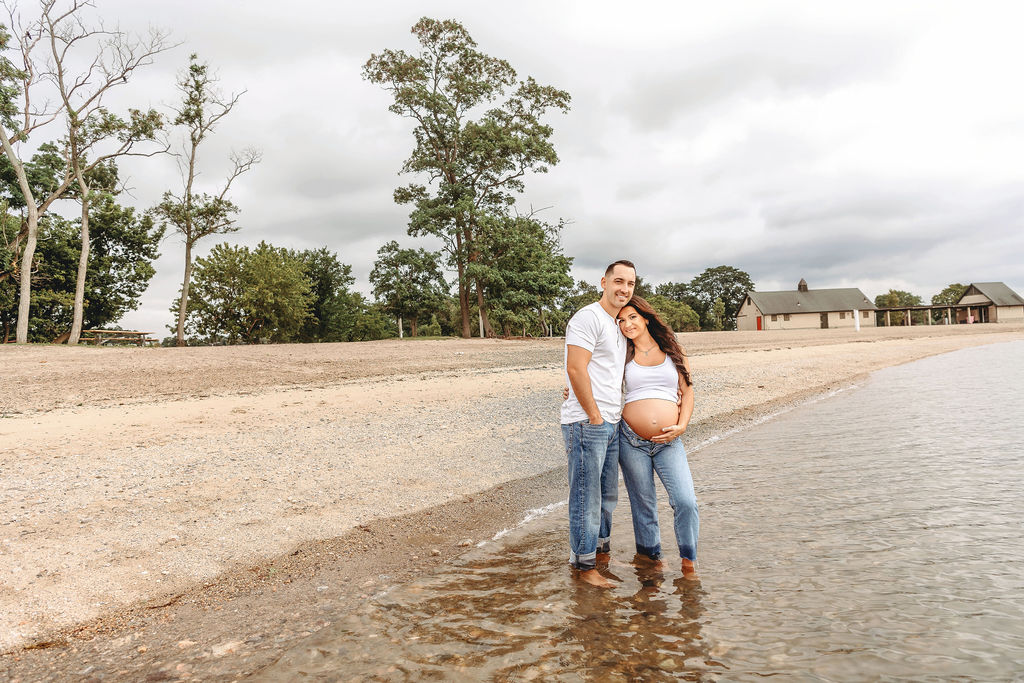A happy expecting couple snuggle while standing in the water in jeans and white shirts while holding the bump after visiting Family Tree Yoga
