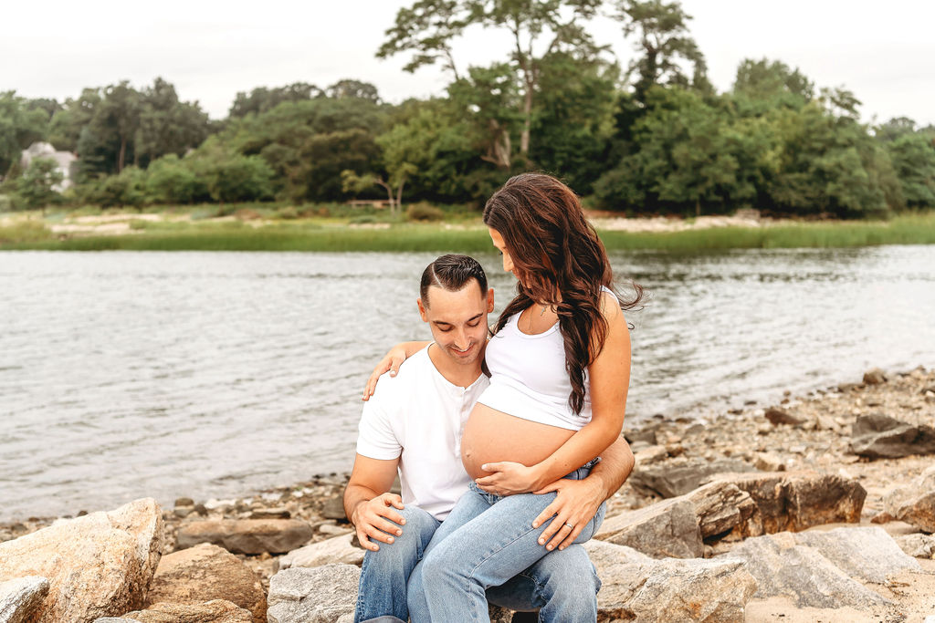 A pregnant woman sits in the lap of her husband with bump out in jeans and white shirts by the water after visiting Family Tree Yoga