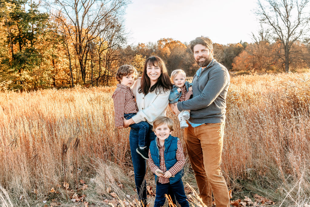 A mom and dad laugh and play with their three toddlers in a field of tall grass at sunset in fall