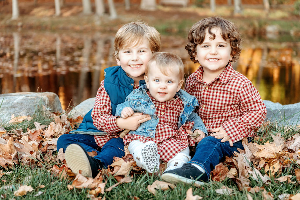 Three toddler siblings sit by a pond in some leaves in matching plaid smiling after meeting nannies in connecticut