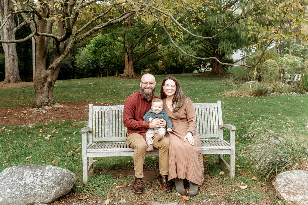 A smiling toddler in a green shirt sits in mom and dads lap on a wooden park bench under a tree in fall