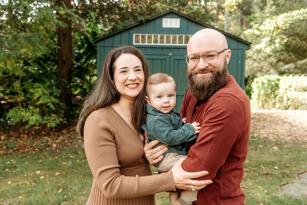 a happy toddler in a green shirt sits in mom and dad's arms in a park as they smile and hug after using connecticut fertility clinics