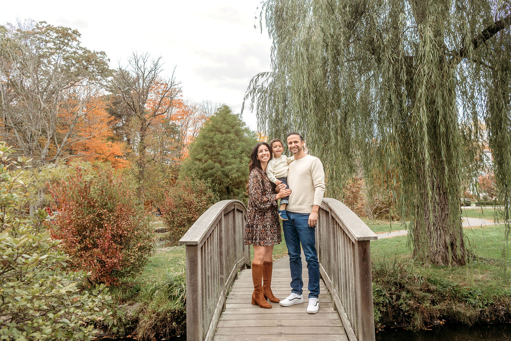 a happy family of three stand smiling on a bridge under a willow tree