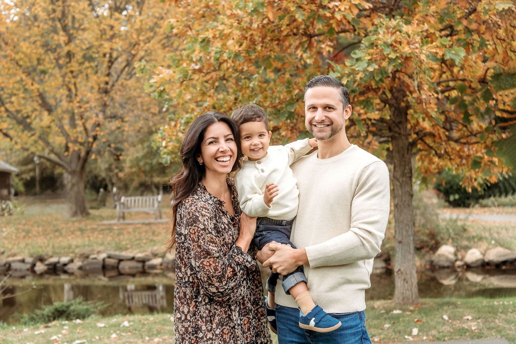 a mom laughs while snuggling her toddler son sitting in dad's arms in a park in fall after visiting a pediatric chiropractor in ct