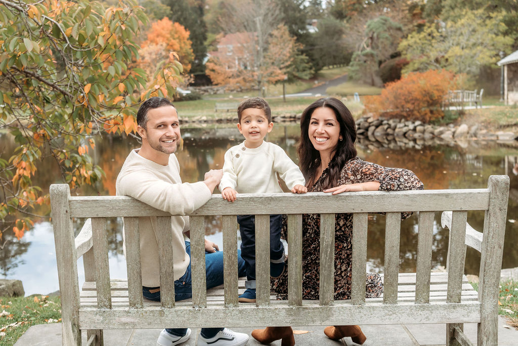 A toddler boy in a white sweater stands on a bench with mom and dad turning around in front of a park pond after visiting a pediatric chiropractor in ct