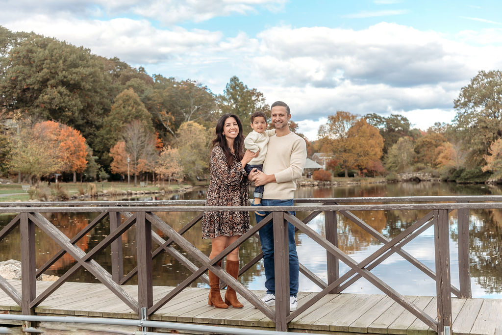 A happy mom and dad stand on a bridge holding their toddler son in fall after visiting a pediatric chiropractor in ct