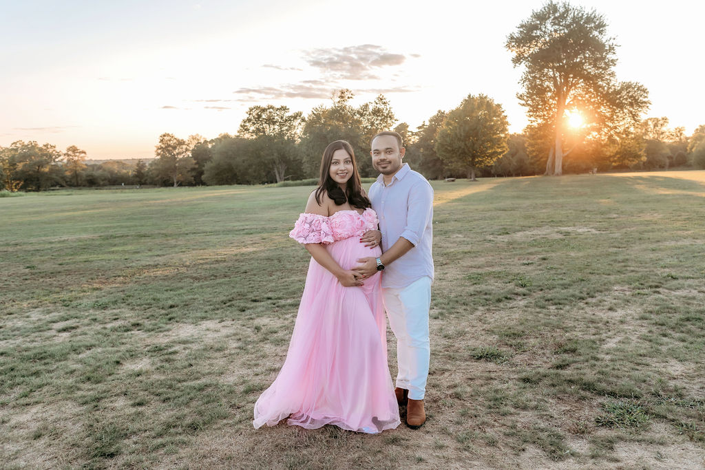 A happy expecting couple stands smiling with hands on the bump at sunset in a park