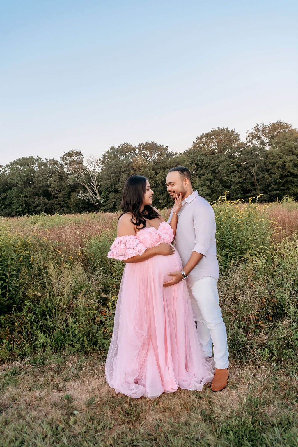 A happy expecting couple snuggle and smile at each other while holding the bump after meeting angela martello in a field of tall grasses