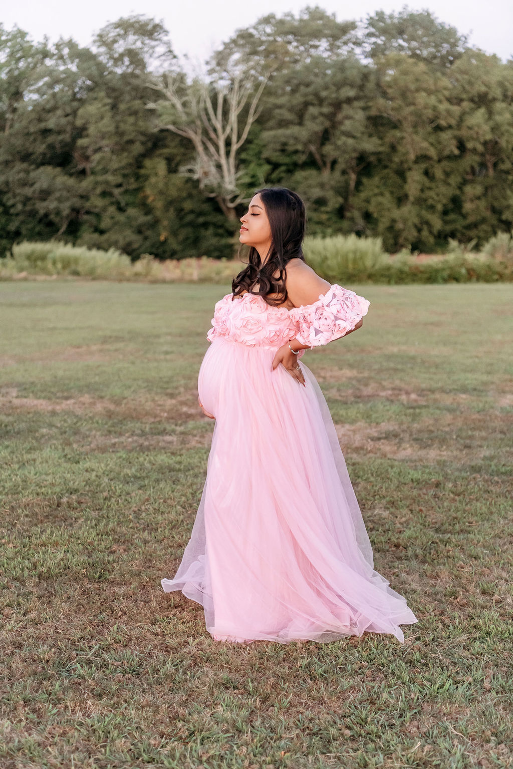 A pregnant woman stands with a hand on her back in a pink maternity gown covered in flowers in a park lawn after meeting angela martello