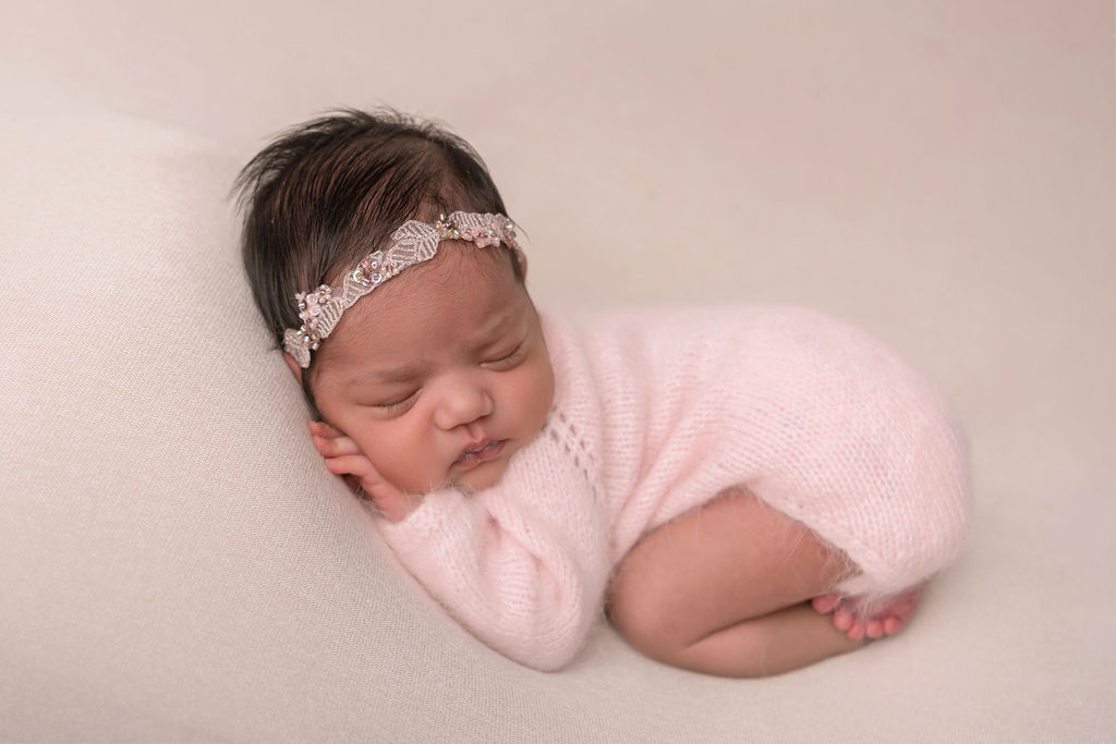 A newborn baby girl sleeps in froggy pose in a pink knit onesie and matching pink bead headband