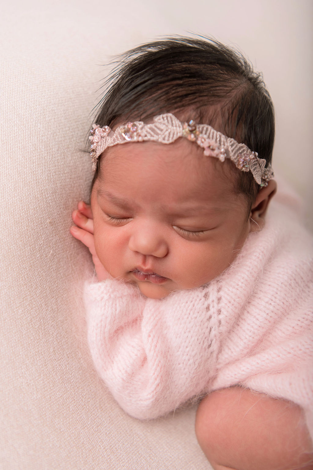 a newborn baby girl in a pink knit onesie and bead headband sleeps on a bed in a studio