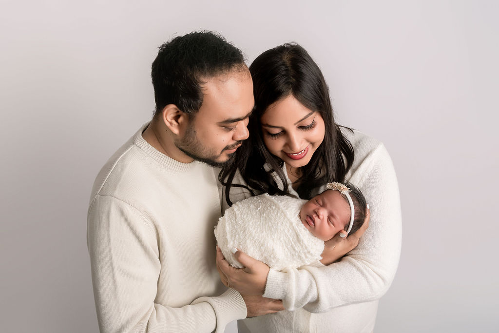 A mother and father in white sweaters gaze down at their newborn cradled against mom's chest while standing in a studio