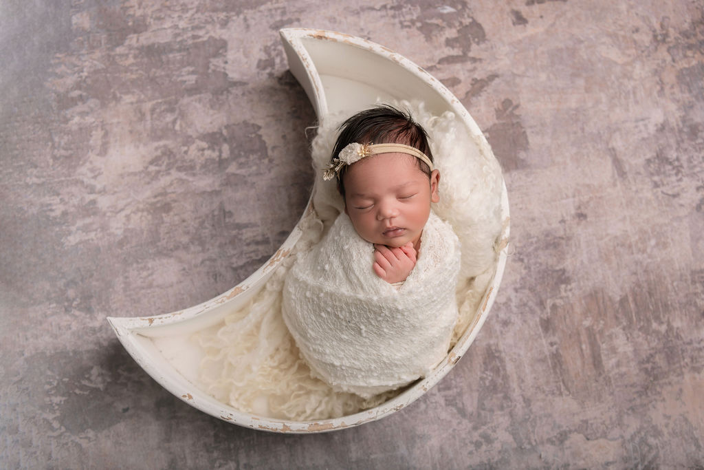 A newborn baby sleeps in a moon shaped bowl in a white swaddle thanks to wee care nanny agency
