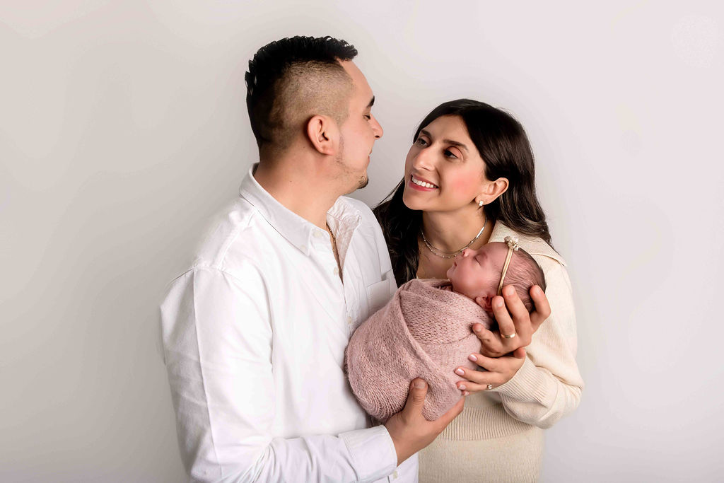 Happy new mom and dad in white and cream stand smiling at each other in a studio holding their sleeping newborn in pink after using baby dash now