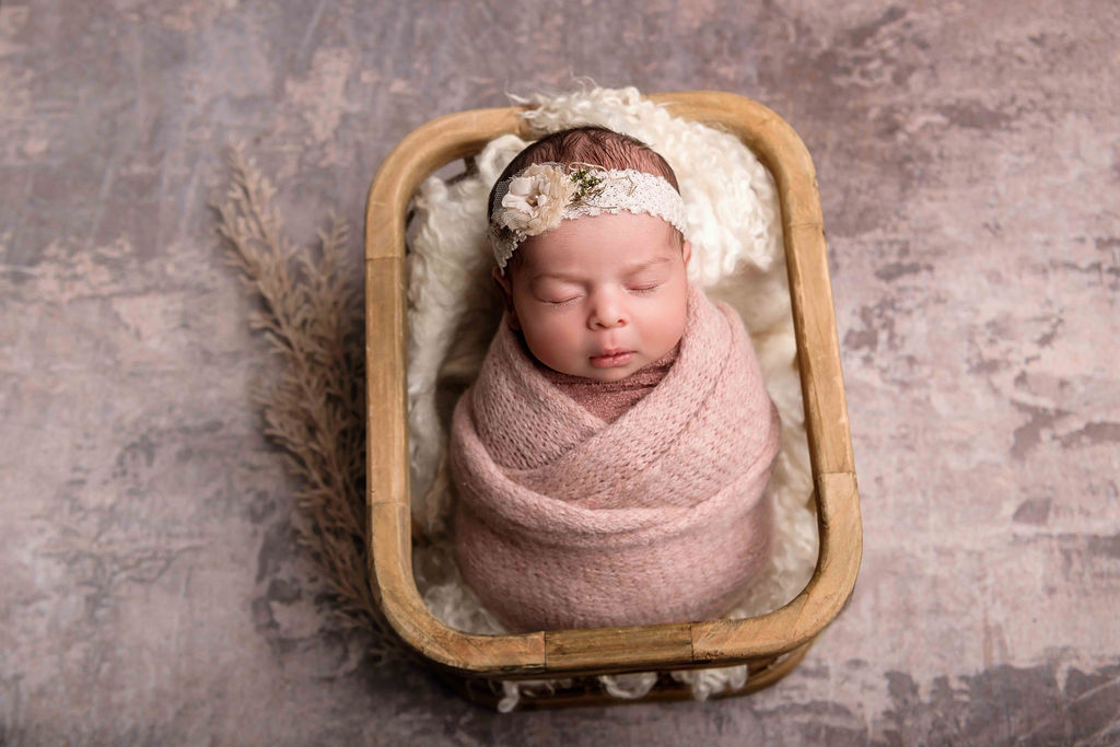 A newborn baby girl sleeps in a headband and pink knit swaddle in a wooden basket