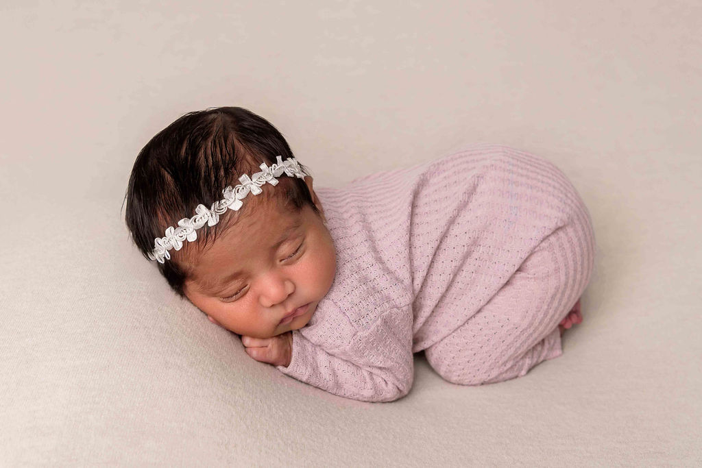 A newborn baby girl in a purple onesie and white headband sleeps in froggy pose